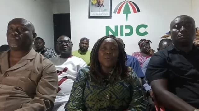 A woman with braids in a patterned green dress speaks at a press conference, surrounded by other individuals. Behind them, the wall features the National Democratic Congress (NDC) logo and a framed portrait of President John Dramani Mahama.