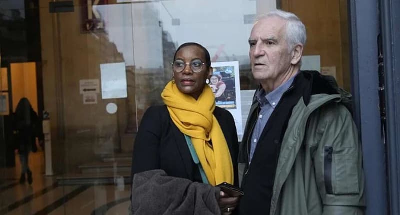 Dafroza Gauthier and her husband Alain Gauthier are pictured standing outside what appears to be a public building with glass doors. Dafroza wears a bright yellow scarf and glasses, while Alain stands beside her in a dark green parka.