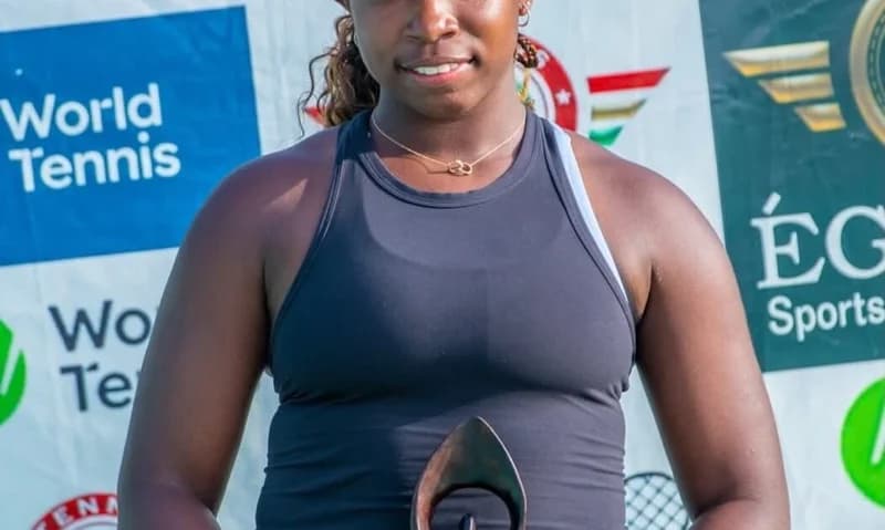 Sydney Wright, a young tennis player, smiles while holding a trophy after being crowned the J100 Accra Champion. She is wearing a black sports top and standing in front of event backdrops featuring World Tennis and ÉG Sports logos.