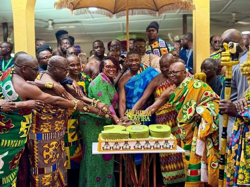 A group of traditional leaders and dignitaries in Ghana gather to cut a large green and gold cake shaped like the number 200. The participants are dressed in vibrant Kente cloth and traditional attire, standing under a ceremonial umbrella to celebrate the bicentenary of a historic Akuapem victory.