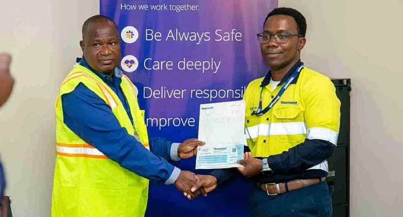 Two men, one in a high-visibility safety vest and the other in a Newmont-branded uniform, shake hands while holding a formal document. The presentation takes place in front of a blue corporate banner highlighting company values.