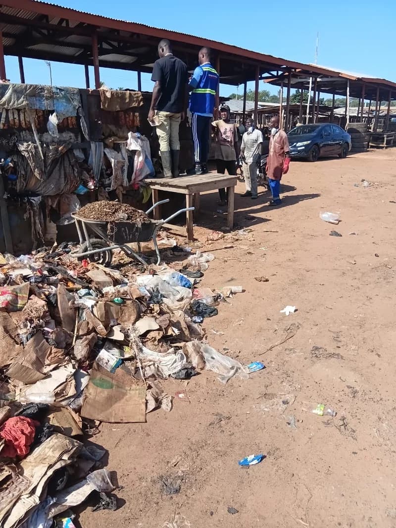 A group of men participate in a sanitation exercise at a market area in Kintampo, Ghana. A large pile of refuse and a wheelbarrow are visible in the foreground, while men in protective gear and reflective vests coordinate the cleanup efforts.