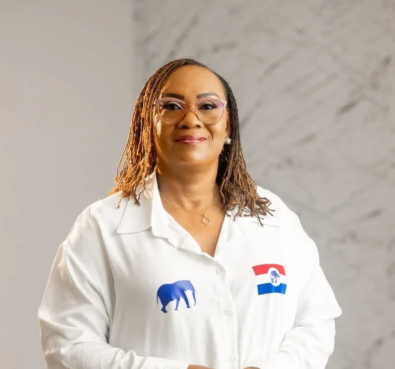 A professional portrait of Mavis Hawa Koomson, a prominent Ghanaian politician. She is smiling and wearing a white button-down shirt adorned with the blue elephant symbol and the red, white, and blue flag logo of the New Patriotic Party (NPP).
