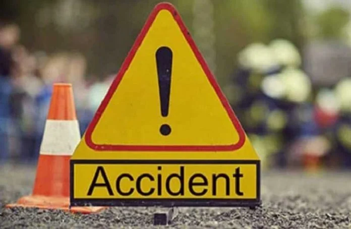 A close-up photograph of a yellow triangular warning sign with a large black exclamation mark, sitting above a rectangular board that reads 'Accident'. In the background, an orange traffic cone and blurred figures of people are visible on a gravel surface, indicating an emergency scene.