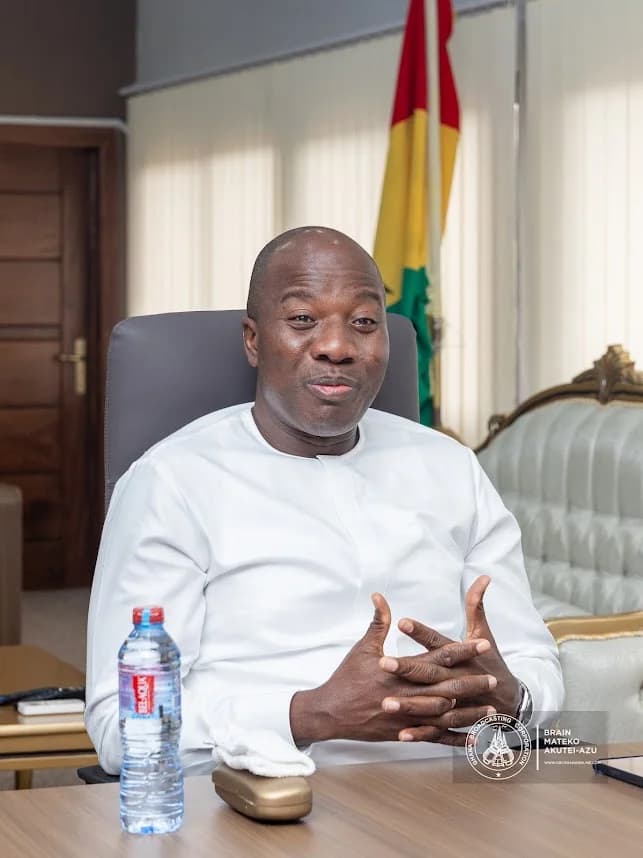 Ghanaian politician Mahama Ayariga is shown seated at a desk in an official office setting, wearing a white traditional garment. The national flag of Ghana is visible in the background as he gestures with his hands during a conversation.
