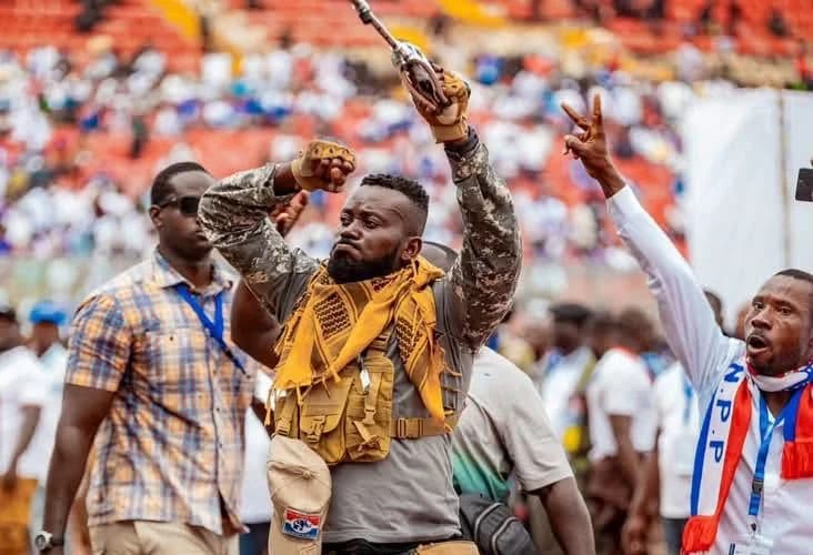 A man in tactical-style gear and a yellow scarf stands with his arms raised in a crowded stadium setting. He is surrounded by other individuals, including one person wearing a scarf with 'N.P.P' text, suggesting a political rally or event for the New Patriotic Party in Ghana.