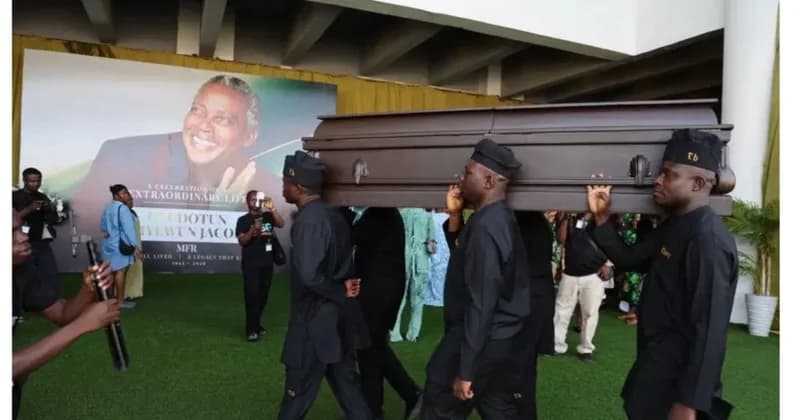 The image depicts the funeral ceremony of the legendary Nigerian actor Olu Jacobs, showing pallbearers in black traditional attire carrying his casket. In the background, a large memorial banner features his portrait and celebratory text, marking the conclusion of his burial rites in Lagos.