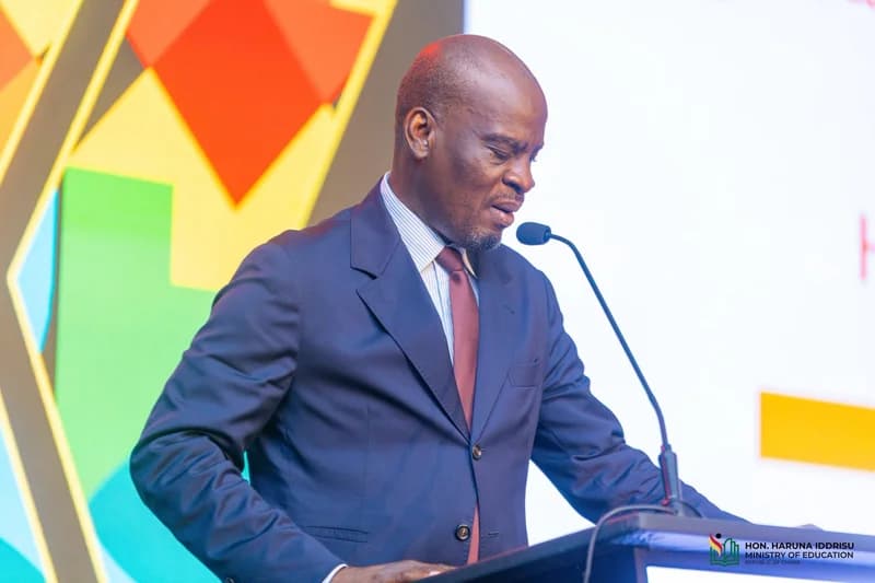 Haruna Iddrisu is captured speaking at a lectern during an official function, likely related to the Ministry of Education. He is dressed in a formal dark suit and tie, appearing focused as he addresses the audience through a microphone.