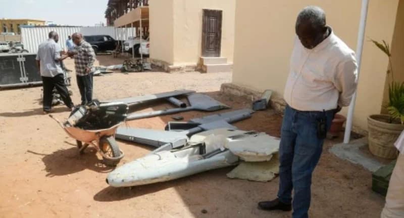 A man in a white shirt and blue jeans stands looking down at the wreckage of a downed drone scattered on the sandy ground of a compound. In the background, other individuals are seen near more debris and parked vehicles, illustrating the physical aftermath of aerial conflict in the region.