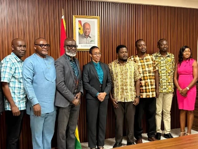 A group of eight individuals, including men and women in formal and traditional attire, pose for a photograph in an office setting. They are standing against a vertical wood-paneled wall that features a portrait of President John Dramani Mahama.