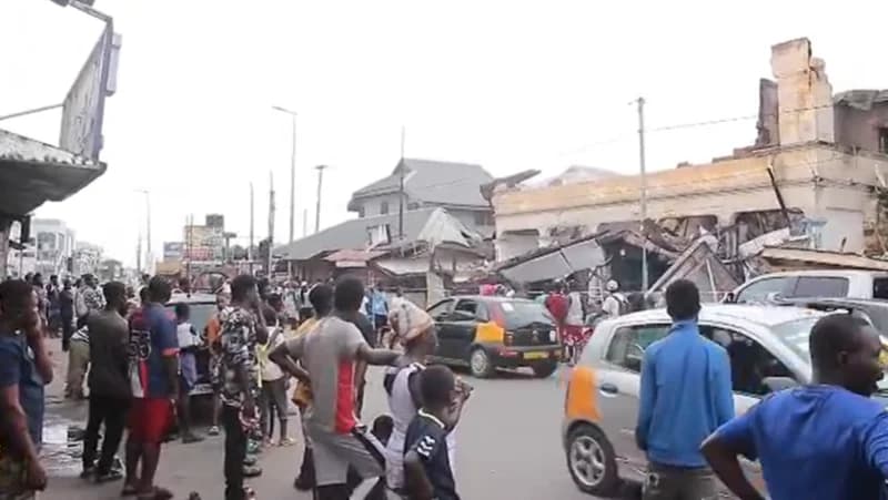 A crowd of people gathers on a street in Koforidua to observe the remains of a building severely damaged by fire. The structural damage is extensive, with collapsed roofing and charred walls visible on the two-story structure situated alongside a busy road with vehicles passing by.