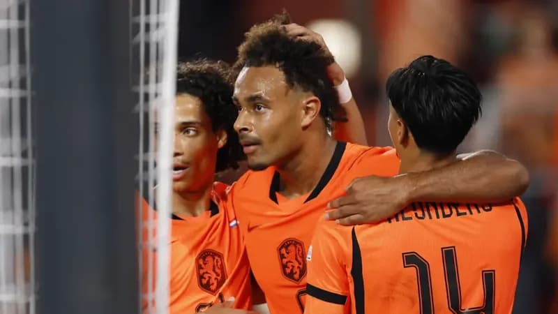 Joshua Zirkzee and his Netherlands teammates, including Tijjani Reijnders, celebrate a goal during an international football match. The players are seen wearing their iconic bright orange kits while embracing near the goal net.