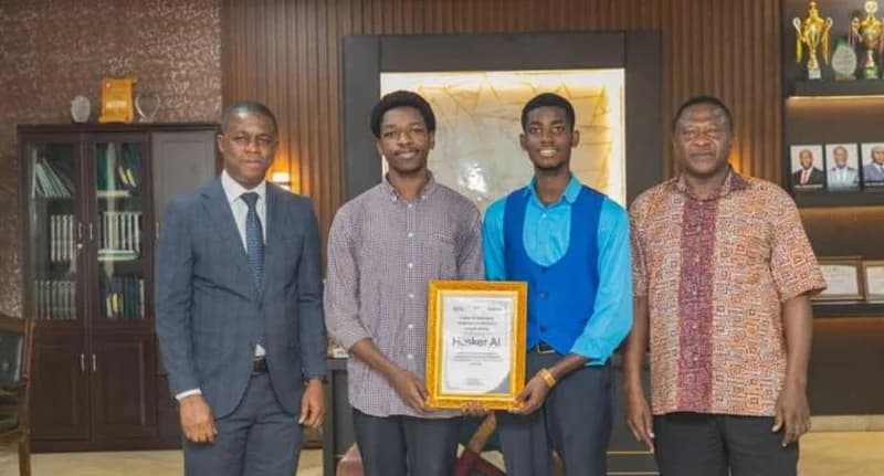 Four men are posed for a photo in an office setting, with the two younger men in the center holding a framed certificate. The group consists of two students and two older officials, likely from KNUST, celebrating a win in a technology competition.