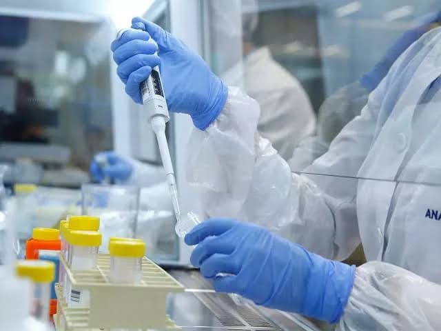 A laboratory technician wearing blue nitrile gloves and a white protective gown uses a precision pipette to transfer liquid into a small sample vial. In the foreground, a rack holds several test tubes with yellow caps within what appears to be a sterile, modern laboratory environment.