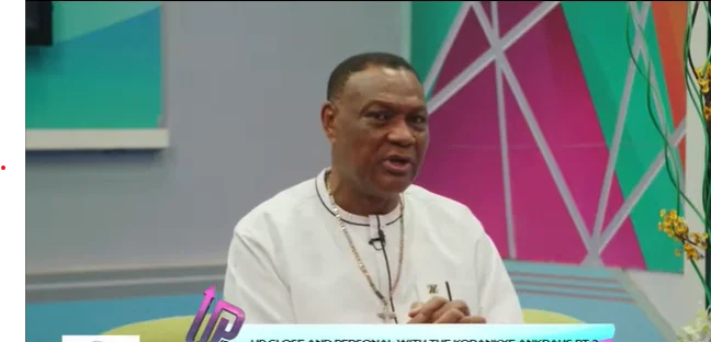 Reverend Sam Korankye Ankrah is captured during a televised interview, speaking directly to the camera. He wears a white traditional tunic and a gold cross necklace against a colorful studio backdrop with geometric patterns.