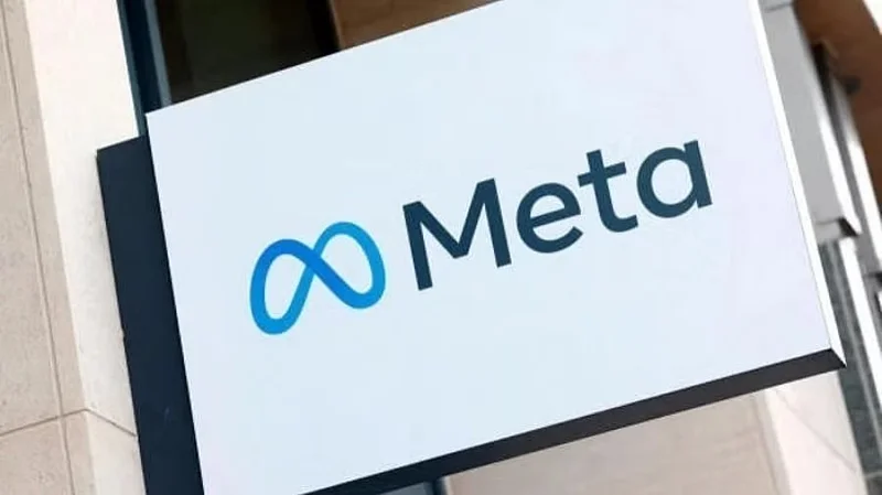 A close-up photograph of a white corporate sign featuring the blue Meta infinity logo and the company name in dark blue text. The rectangular sign is mounted on the exterior of a modern building with light-colored masonry.