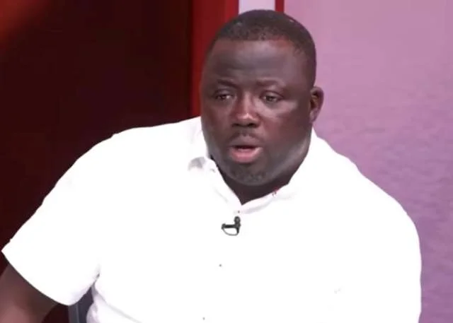 A portrait of Mustapha Gbande, the Deputy General Secretary of the National Democratic Congress (NDC), captured during a televised interview. He is wearing a white button-down shirt with a small lapel microphone attached, set against a dark studio background.