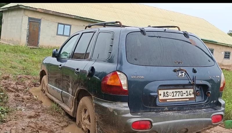 A dark blue Hyundai Santa Fe SUV is stuck in thick mud in a rural setting in Tamale. The vehicle features a Ghanaian license plate (AS 1770-14) and is parked near a simple building with a corrugated metal roof.