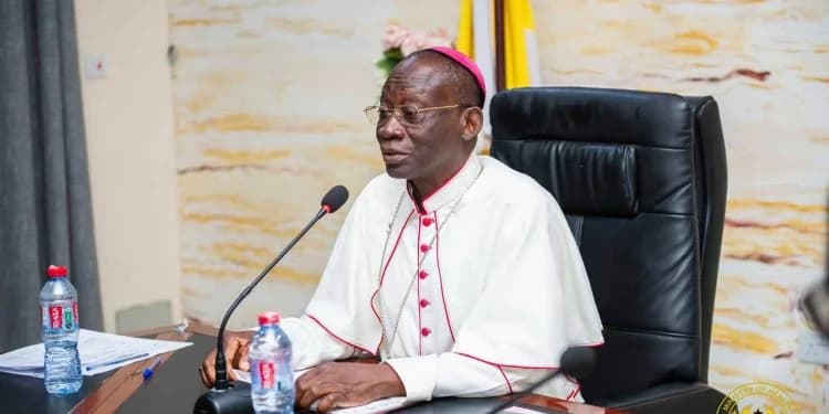 Most Rev. Matthew Kwasi Gyamfi, President of the Ghana Catholic Bishops' Conference, is seen speaking into a microphone during a meeting. He wears a white cassock with red accents and a magenta skullcap, seated at a desk with bottled water and a Ministry of Health logo visible in the corner.