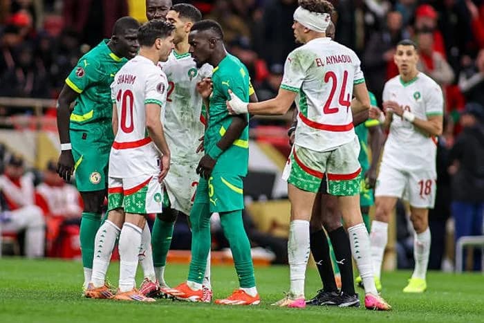 Football players from Senegal and Morocco are seen in a heated confrontation on the pitch during a match. The scene captures players in green and white kits arguing closely, with Morocco's Brahim Diaz and a bandaged El Aynaoui prominently featured amid the tension.