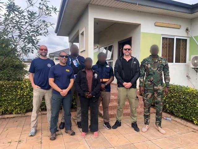A group of seven law enforcement and military personnel stand together in front of a building in Ghana. The group includes individuals wearing FBI-branded t-shirts, INTERPOL tactical vests, and one person in a Ghana Armed Forces camouflage uniform, signifying a joint international operation.
