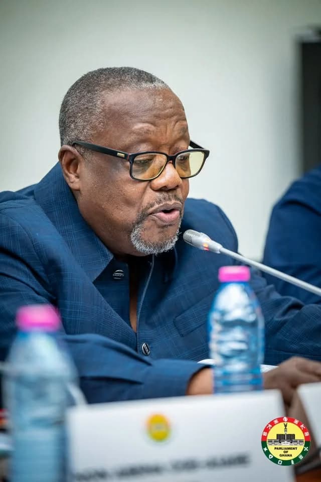 Hon. James Klutse Avedzi, Chairman of the Public Accounts Committee (PAC), is seen speaking into a microphone during a parliamentary hearing. He is wearing a blue patterned suit and glasses, with a Parliament of Ghana logo visible in the bottom right corner.