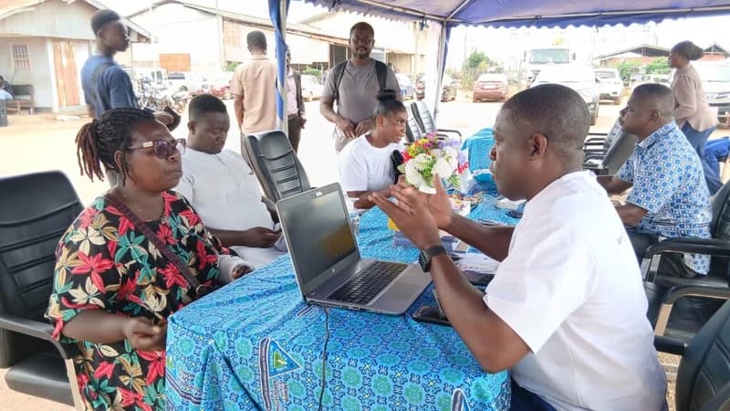 A group of people are gathered under a blue canopy for what appears to be a community outreach or customer service engagement. A man in a white shirt is seen speaking to a woman at a table equipped with a laptop, while other individuals look on or wait in the background. The setup features a blue patterned tablecloth with logos related to the Volta River Authority (VRA).