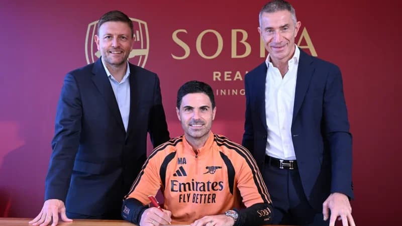 Arsenal manager Mikel Arteta is seen signing a contract extension while flanked by club executives Richard Garlick and Edu Gaspar. Arteta is wearing an orange training kit, and they are positioned in front of a maroon wall featuring the club's crest and the Sobha Realty Training Centre logo.