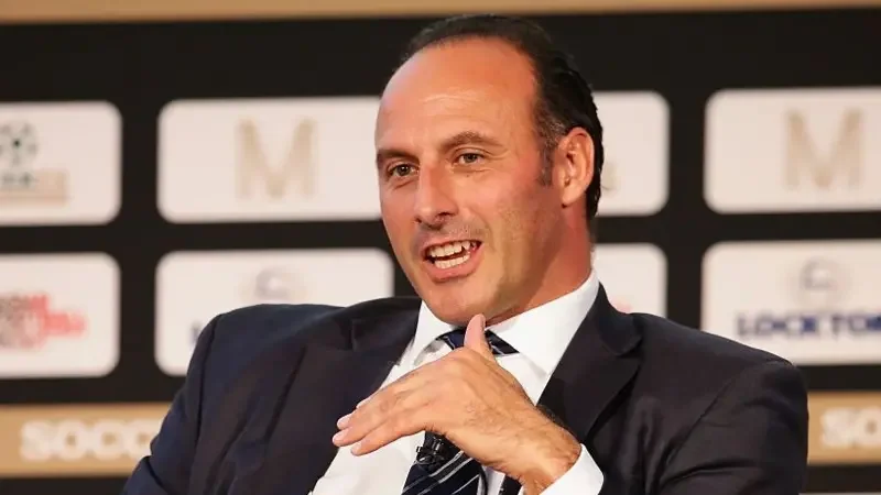 A medium close-up photograph of former footballer Ramon Vega speaking at a public event. He is dressed in professional attire, including a dark suit and striped tie, while gesturing with his hand against a branded backdrop featuring various logos.