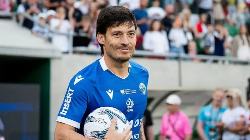 Former Manchester City midfielder David Silva is captured smiling while walking on a football field holding a soccer ball. He is wearing a blue athletic kit with several logos, set against a background of blurred spectators in a stadium setting.