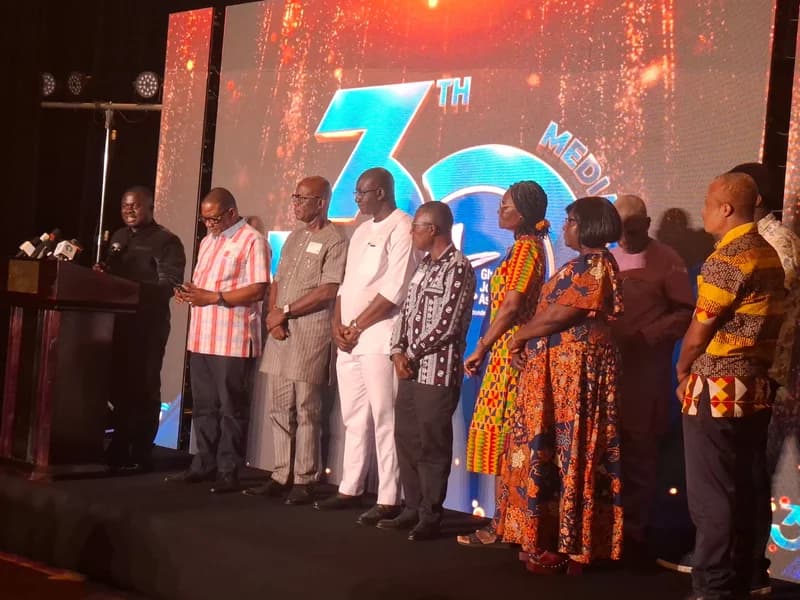 A group of individuals, including leadership from the Ghana Journalists Association (GJA), stand on a stage during a formal media event. Albert Kwabena Dwumfour, the GJA President, is pictured speaking at a podium with several microphones, while other members stand in a line behind him against a large digital screen backdrop.