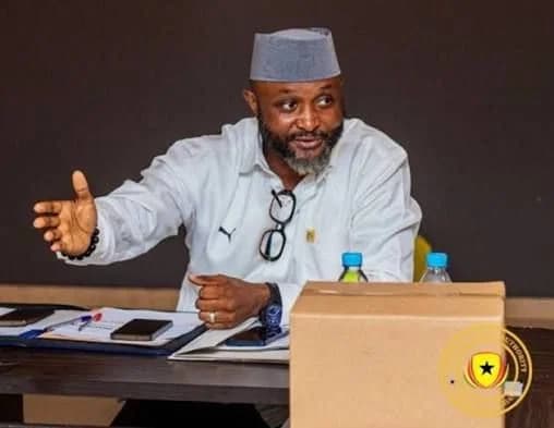 A photograph of Ghanaian sports journalist Yaw Ampofo Ankrah seated at a table, gesturing with his hand during what appears to be a formal meeting or briefing. He is wearing a grey cap and a white shirt with glasses hanging from his collar, with documents, phones, and water bottles visible on the desk.