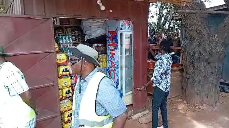 A roadside kiosk stocked with beverages and snacks is being inspected, with a man in a high-visibility safety vest and wide-brimmed hat standing in the foreground. The shop appears to be located on a pedestrian path or roadside area near a large tree, typical of informal trading spots in Accra.