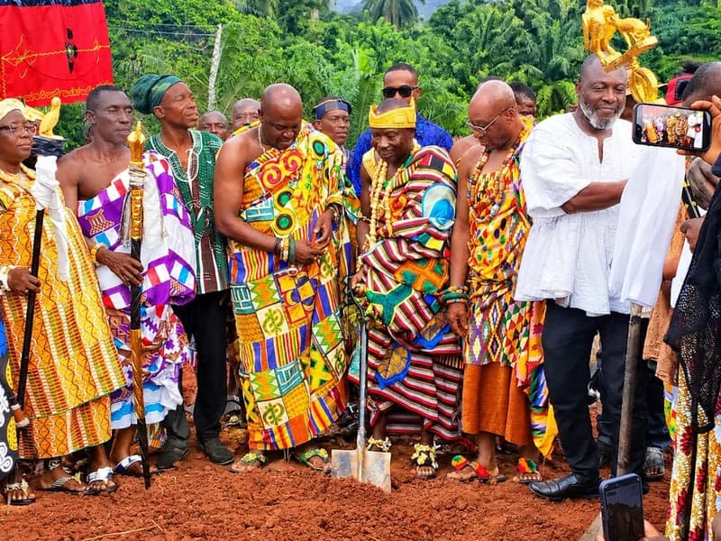 Togbega Sessinu VI, dressed in vibrant traditional kente cloth, performs a ground-breaking ceremony by digging into the red earth with a shovel. He is surrounded by other traditional leaders, community members, and onlookers in a lush, green rural setting in Ghana.