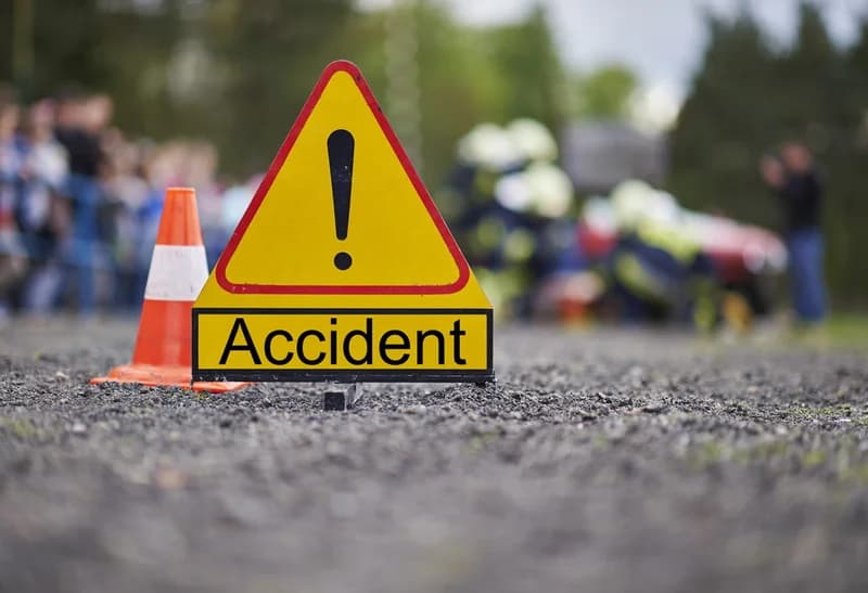 A yellow triangular warning sign with an exclamation mark and the word "Accident" sits prominently on a gravel road. In the blurred background, emergency responders and a crowd are visible at the scene of an incident, accompanied by an orange and white traffic cone.