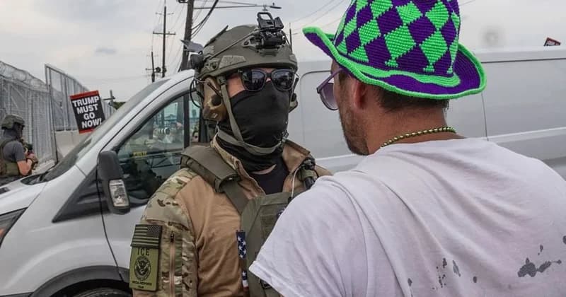 An Immigration and Customs Enforcement (ICE) officer in full tactical gear and a face mask stands face-to-face with a civilian wearing a colorful purple and green hat. The officer's uniform bears an ICE Special Response Team patch, while a protest sign in the background reads "TRUMP MUST GO NOW!" near a white van and security fencing.