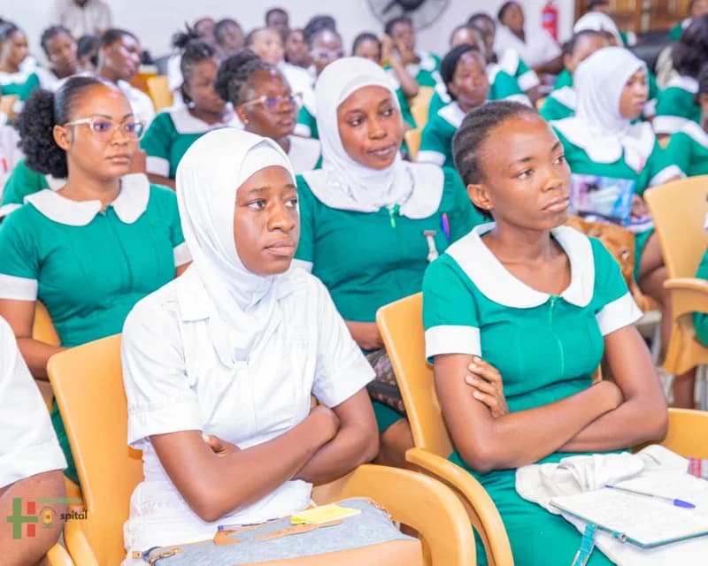 A group of female nursing students sitting in a lecture hall or assembly, appearing attentive. Most students are wearing green uniforms with white collars, while one student in the foreground is dressed in a white uniform and a white hijab.