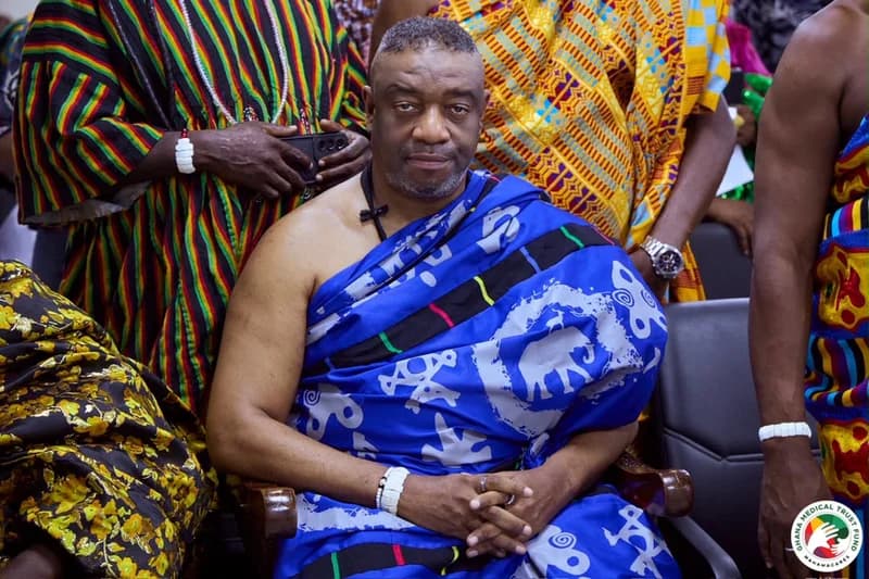 King Tackie Teiko Tsuru II, the Ga Mantse, is captured seated in vibrant blue traditional regalia adorned with elephant and adinkra motifs. He is surrounded by attendants in colorful Kente and traditional smocks, during an event likely related to the Ghana Medical Trust Fund.