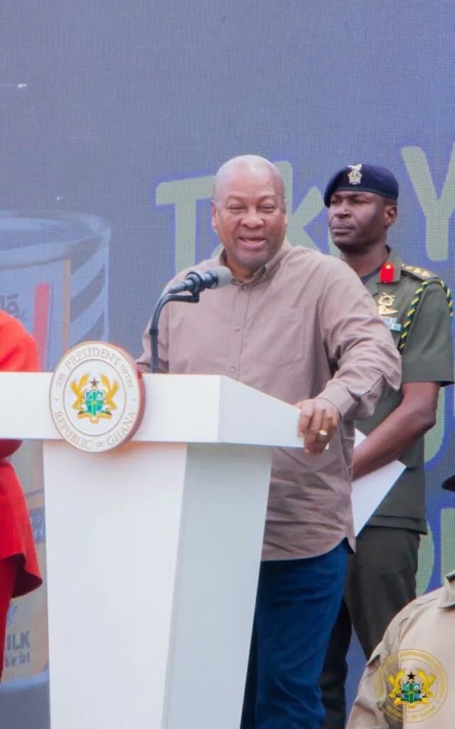 President John Dramani Mahama stands at a white podium bearing the Presidential Seal of Ghana during an official event. He is dressed in a brown button-down shirt and is accompanied by a military aide-de-camp standing behind him against a digital background.