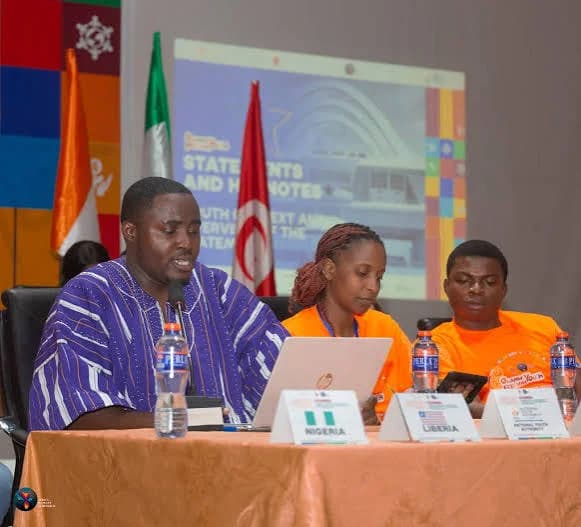 Three delegates are seated at a conference table during the Africa We Want Symposium in Accra. A man in a traditional Ghanaian blue and white smock sits beside two colleagues in orange t-shirts, with placards for Nigeria, Liberia, and the National Youth Authority positioned in front of them.