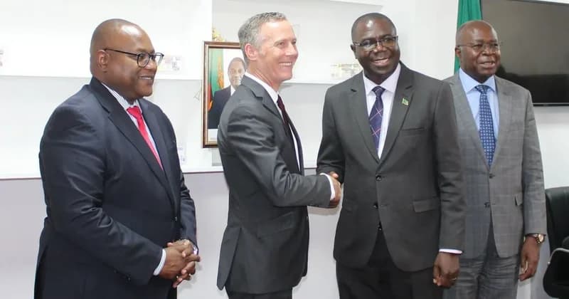 A photograph showing four men in formal suits during a diplomatic meeting. Two men in the center are shaking hands, while a portrait of Zambian President Hakainde Hichilema is visible in the background, signifying an official government agreement.