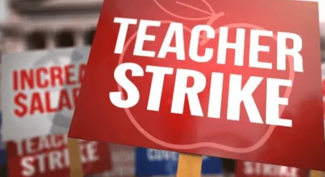 A close-up photograph of several protest placards held during a demonstration. The primary red sign in focus displays the words 'TEACHER STRIKE' in bold white capital letters over an apple graphic, while blurred background signs demand salary increases.
