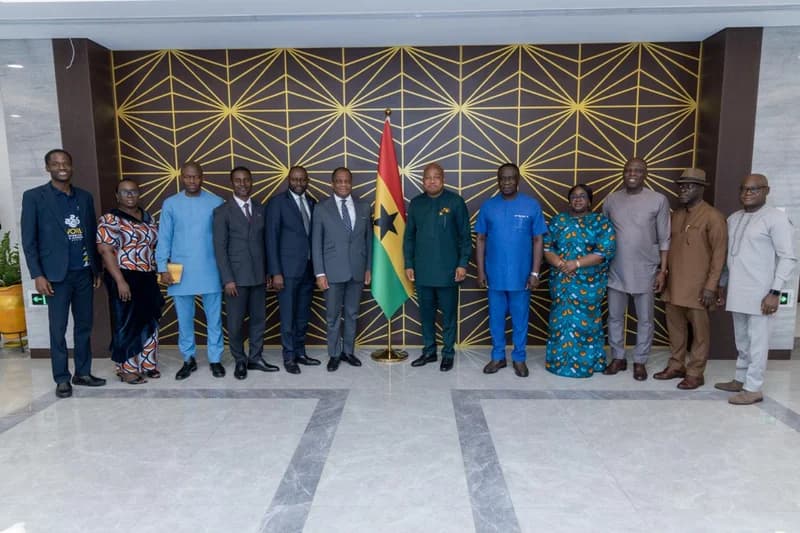 A group of twelve individuals, including religious leaders and government officials, pose for a formal photograph in a modern office setting. Centered in front of a Ghanaian flag and a gold geometric patterned wall, the delegation from the Assemblies of God World Congress meets with political figures. The attendees are dressed in a variety of formal suits and traditional Ghanaian smocks and dresses.
