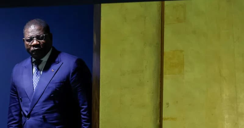 A portrait of President Umaro Sissoco Embaló of Guinea-Bissau, wearing a dark blue double-breasted suit and glasses. He stands with a serious expression against a background of deep blue and a textured gold-colored wall.