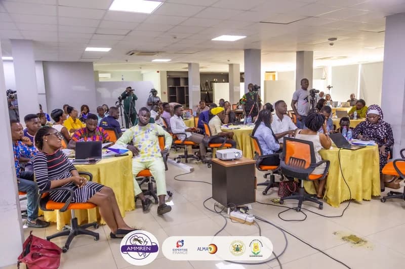 A group of journalists and professionals are seated at round tables during a workshop or media engagement session in an office setting. Participants are seen using laptops and taking notes, while professional cameras and a projector indicate an official presentation or training event.