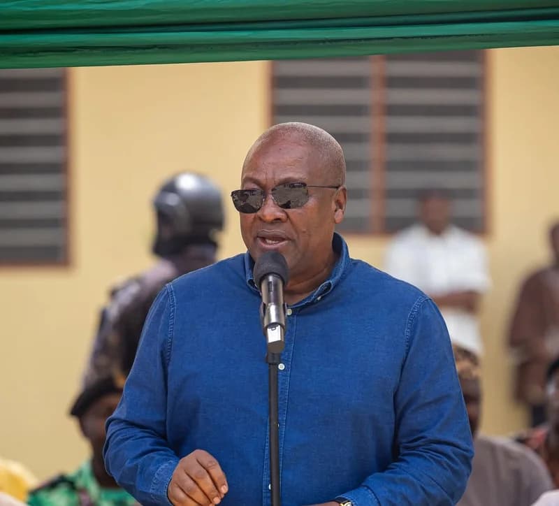 President John Dramani Mahama is pictured speaking at a public gathering while wearing a blue denim shirt and sunglasses. He stands behind a microphone, addressing an audience against a backdrop of a building and security personnel.