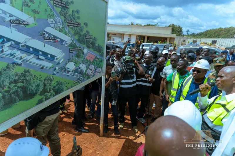 President John Mahama, wearing a white hard hat and yellow safety vest, inspects a detailed architectural site plan for the Bechem Poultry and Meat Processing Factory. He is surrounded by a large contingent of officials, security personnel, and media members who are documenting the site visit.