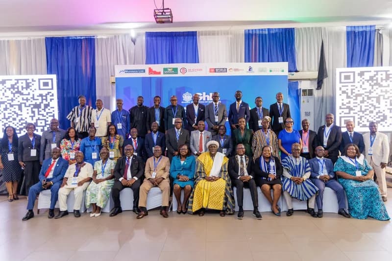 A large group of professionals, including traditional leaders, are posing for a group photograph at a Ghana Institution of Surveyors (GhIS) event. The attendees are dressed in formal business suits and traditional Ghanaian cloths like kente and fugu, seated and standing in front of a blue backdrop featuring various corporate sponsor logos.