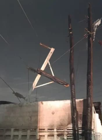 A photograph showing severely burnt utility poles and damaged electrical infrastructure following a fire incident. The wooden poles and cross-arms are charred, with power lines sagging precariously against a dark, overcast sky.