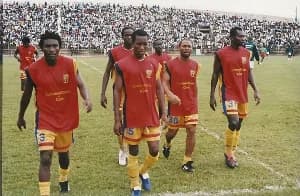 A group of Accra Hearts of Oak football players walk across a pitch in their iconic red, yellow, and blue kits. In the background, a large crowd of spectators fills the stadium stands, capturing a moment from a past era of the Ghana Premier League.